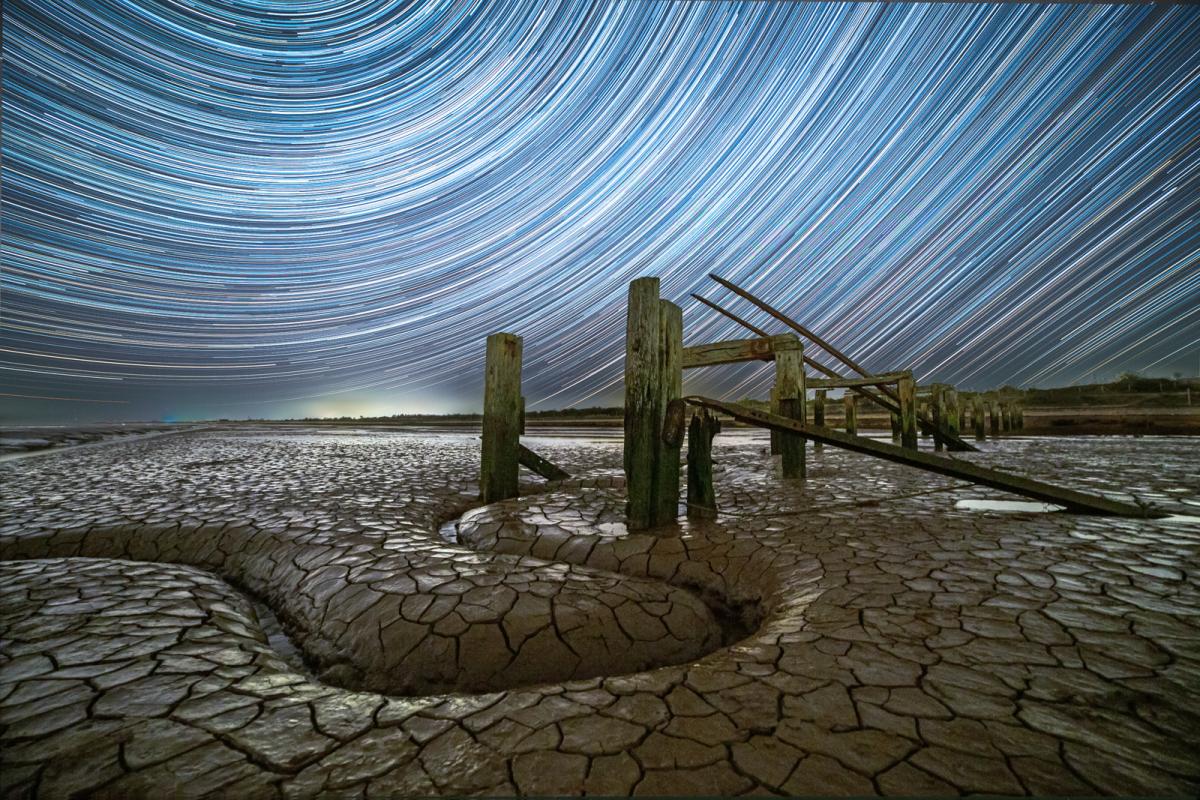 Image of dry mud terrain where the sea is out, with a curved path through the landscape with water in and big cracks in the mud. Rotted wooden sea defenses come out from the land. Behind in the blue sky are star trails forming concentric curved lines 