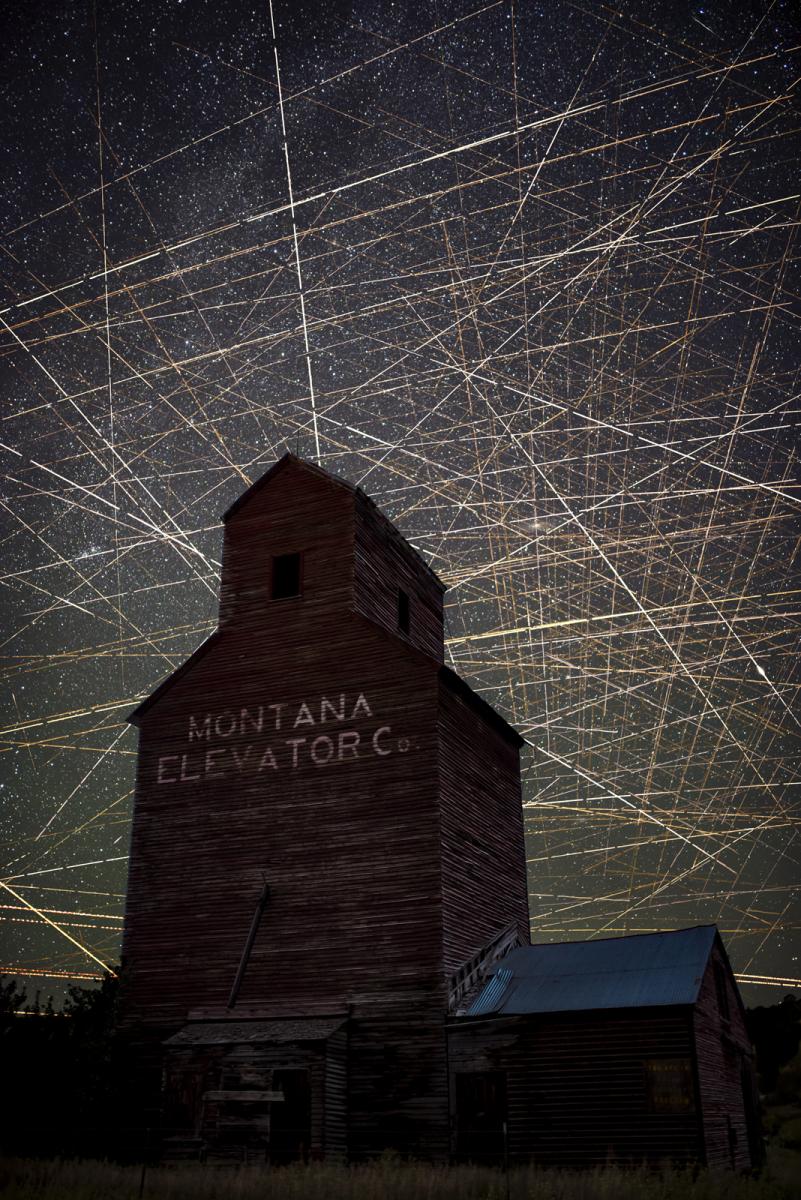 Image showing an old wooden mill style building with Montana elevator written on the side. In the sky behind is a very dense long exposure of satellite trails, which criscross and zigzag across the sky to form hundreds of interacting straight lines.