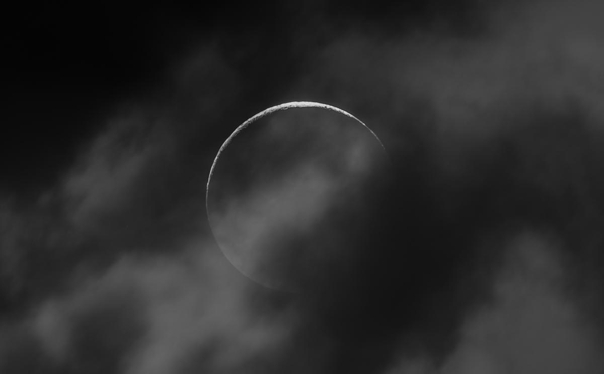 Image of a dark black sky with wispy grey clouds, in the middle of the image is the Moon which appears as a dark black sphere with only a very thin sliver of the top curve illuminated by sunlight, the sliver that is lit shows a cratered and textured surface. A portion of wispy grey cloud covers the dark part of the Moon