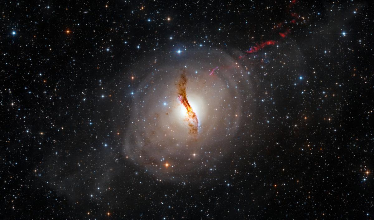 Image of a very dark starry sky with hundreds of stars in a range of sizes and colours including orange, blue and white. At the centre of the image is a round cloud of white dust which is slightly translucent. At the centre of the orb is a yellow/brown core which is a vertical wavy shape. In the upper right of the image is a streak of thick red clouds of gas