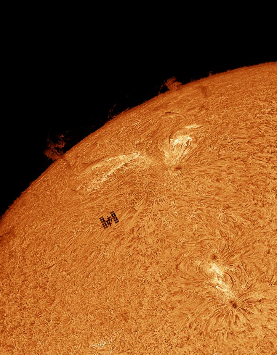 Image showing a section of the Sun, with a curved edge, whose surface is made up of swirling orange patches. Silhouetted in almost the middle of the image is the H shape of the International Space Station and its solar panels