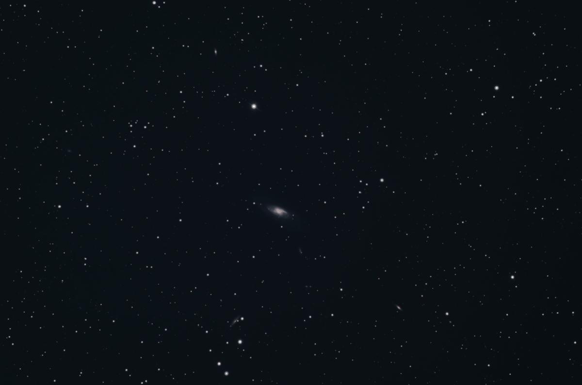 Image showing a dark black sky with multiple stars dotted around, in the centre of the image is a distant white oval shaped smudge which is a galaxy M106, of which it is possible to make out a dark white core with blue and purple spiral arms coming off of it