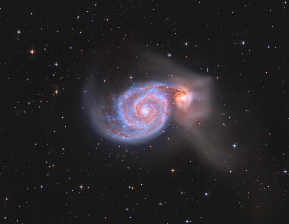 Image of a spiral galaxy in bright light blues, reds and purples. Trails of white haze are coming off of the galaxy and to its right is a small pink round shape surrounded by haze. Behind it is a black sky with stars dotted around the image.