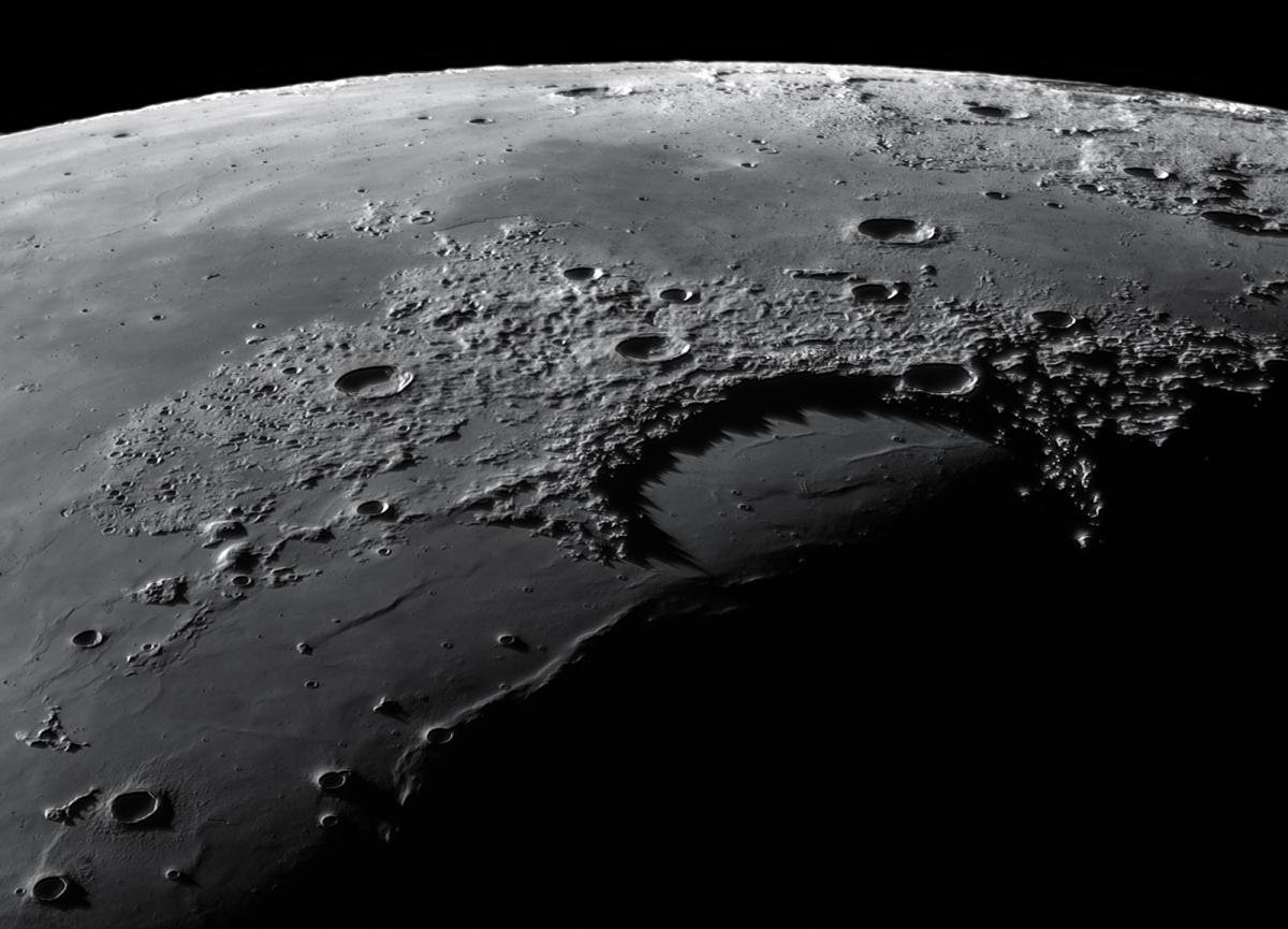 Image showing a close up of the surface of the moon from a diagonal angle looking down from just above it, with the curve of the moon visible in the top of the image. The lunar surface is pockmarked with many round craters of different sizes and depths, and raised areas showing hills and mountains on the surface. The image fades to blackness in the bottom right hand corner.