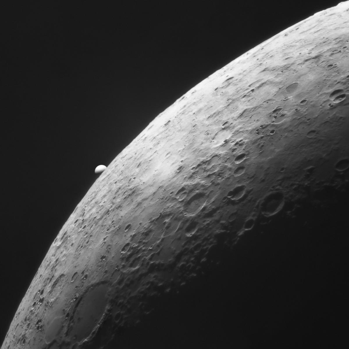 Image showing a close up of a curved portion of the Moon, with many craters visible on the surface. Peeking out from behind the moon is the round shape of Venus, of which the top half is visible and the bottom part is in darkness.