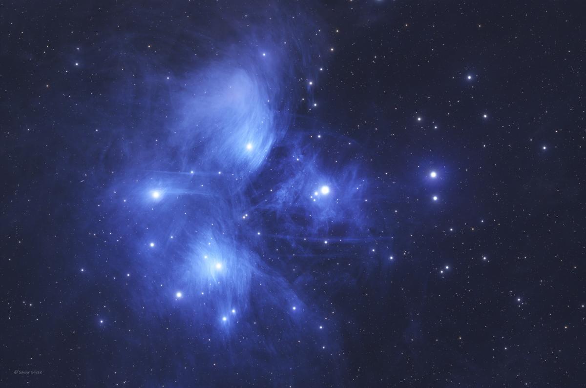 Close-up telescope view of the Pleiades star cluster, showing a series of bright white-blue stars surrounded by a haze of dark blue light