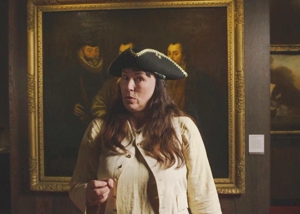 A woman wearing a captain's hat and light cream top looks directly at the camera, with an old portrait in the background