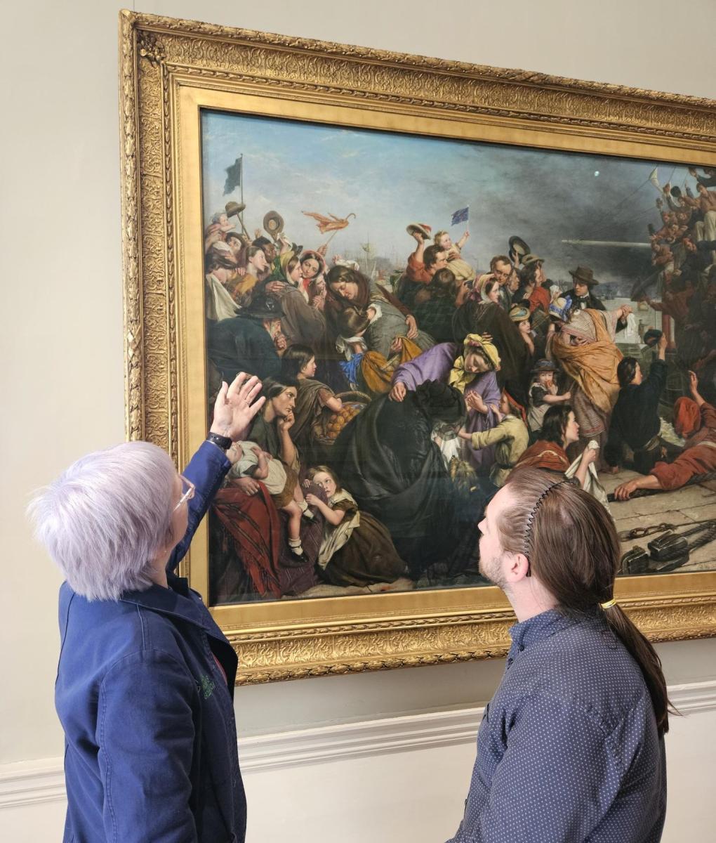 Photo of a woman wearing a blue uniform gesturing to a large painting on a wall in a gold frame, depicting lots of people. A man stands on the right side of the image looking up at the painting