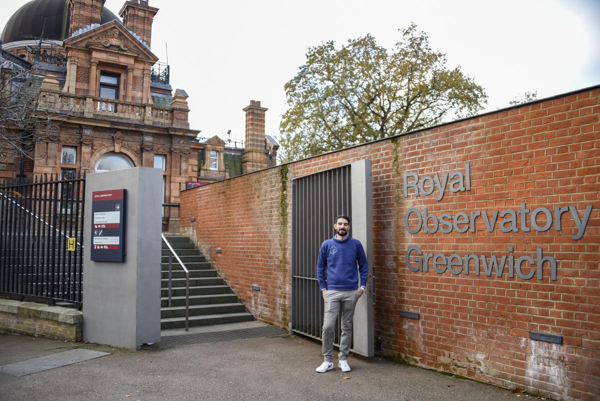 A member of staff at Royal Museums Greenwich stands at the entrance to the Royal Observatory. He is wearing a blue jumper, and behind him a set of stairs leads up to the Observatory building