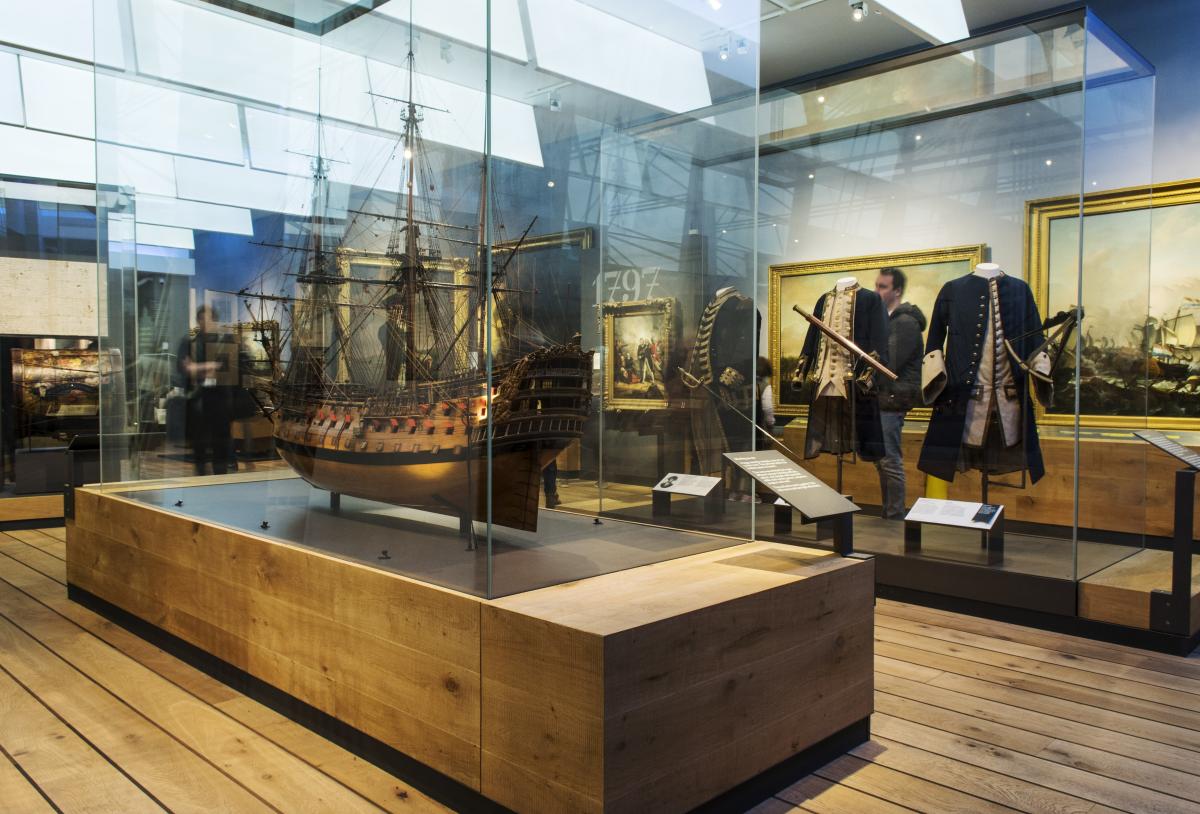 The Nelson, Navy, Nation gallery at the National Maritime Museum. A ship model in a glass case is in the foreground, with naval uniforms and oil paintings in the background