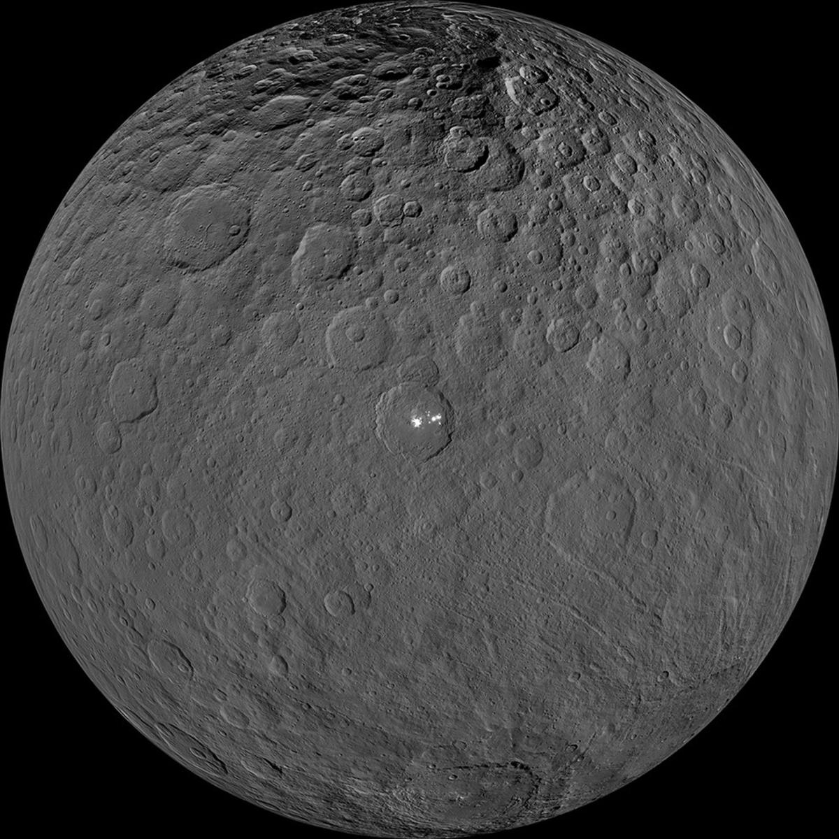 Photograph of round grey dwarf planet Ceres with cratered uneven surface with a white spot in the middle