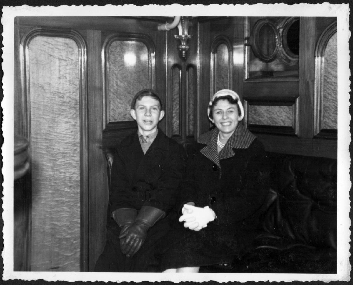 Black and white photograph of a boy with his mother, smiling brightly. They are sat in a wood-panelled saloon: the boy wears large black leather gloves and a woollen hat, the mother white gloves, pearls and a white hat