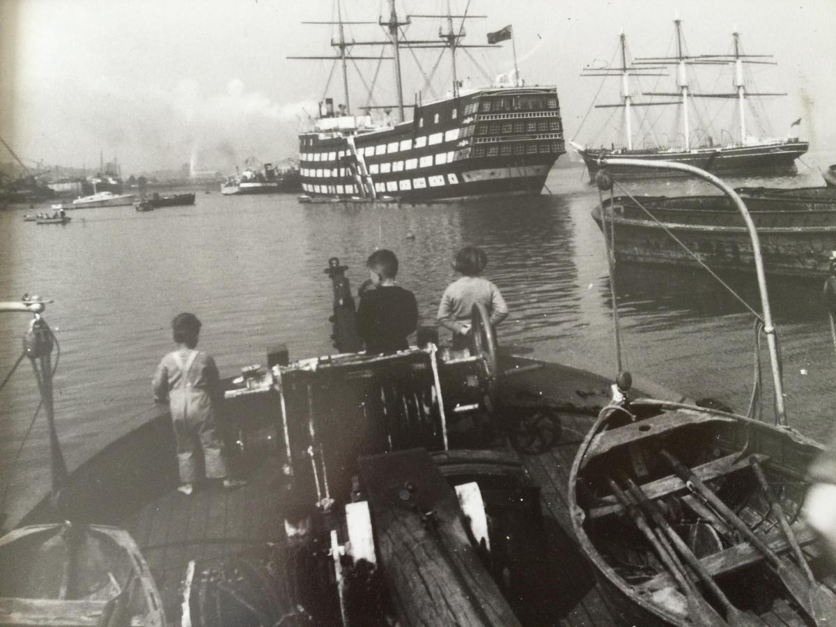 Black and white photograph showing three children standing at the stern of a boat, looking out at the River Thames. In the distance the large training ship HMS Worcester is at anchor, and to the right the tea clipper Cutty Sark