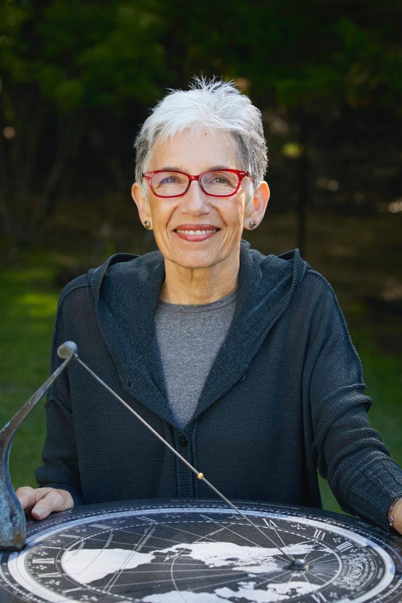 Headshot of Dava Sobel; a woman with short hair and red glasses wearing a black and grey