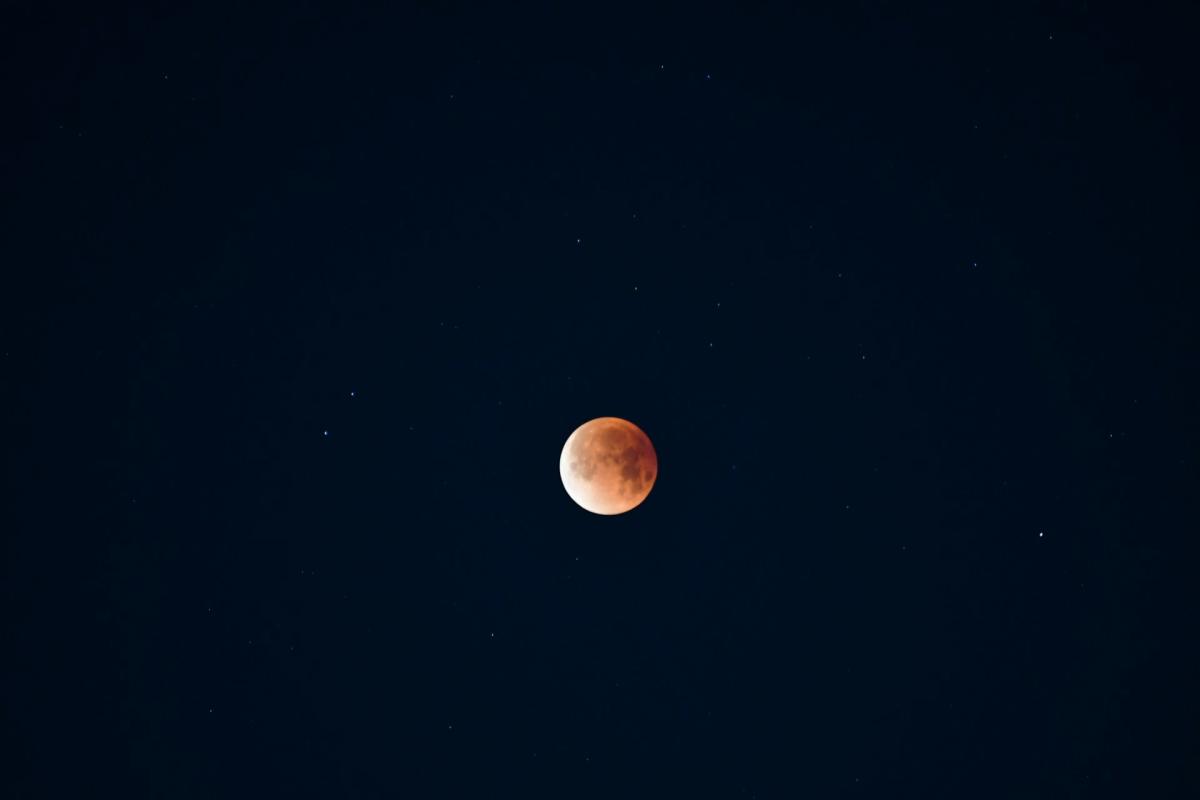 Photo of the Moon which is 90% illuminated in a rust red hue against a starry black sky