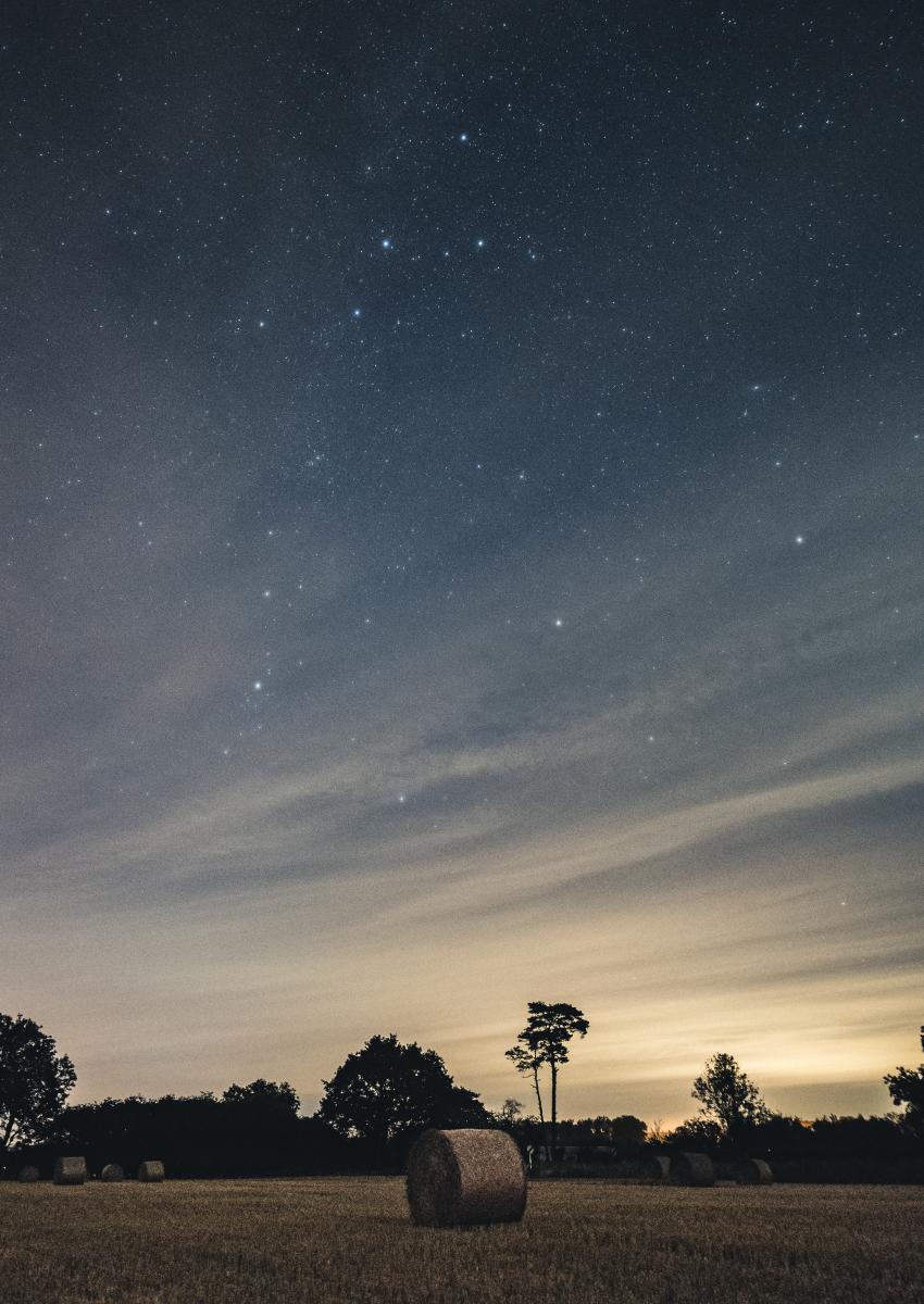 Photo of sky at twilights with stars in upper sky, with clouds on horizon that are lit slightly by the Sun. The foreground is a hay field with a hay bale