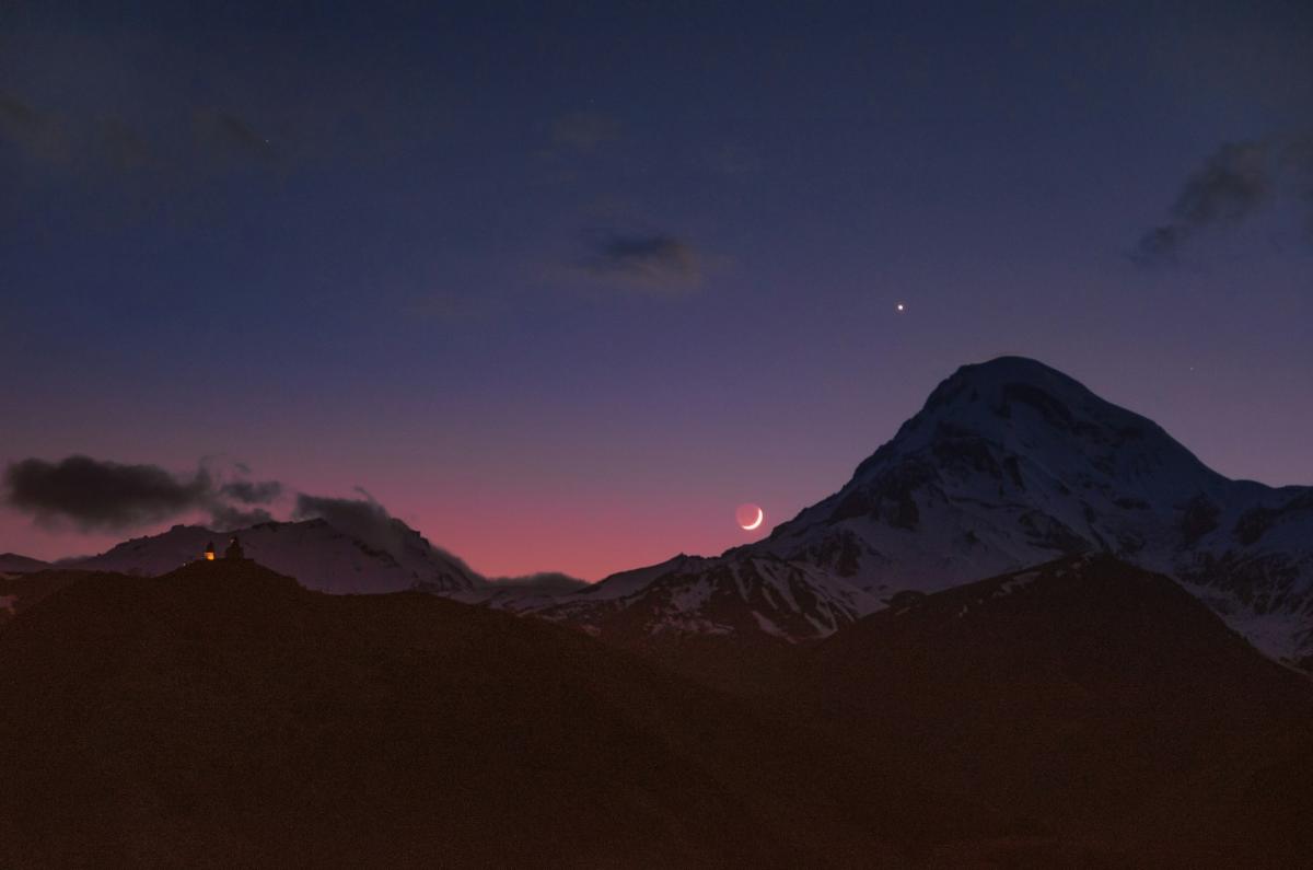 Mountainous landscape with mountains slightly silhouetted in foreground, with pink and purple sunset in sky, towards the horizon is a crescent moon and slightly further up is Venus, a bright white point of light