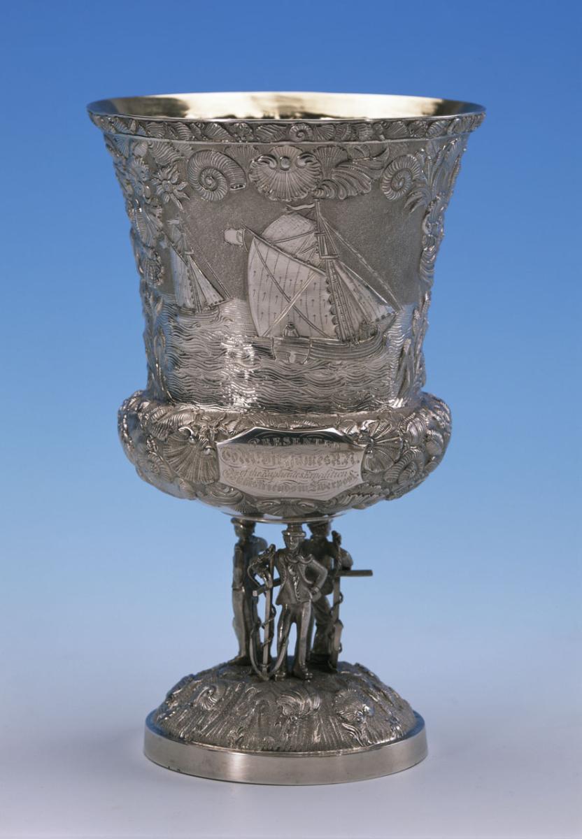 Photo of a silver cup on a stand with finely engraved decorations of ships on the sea and shells