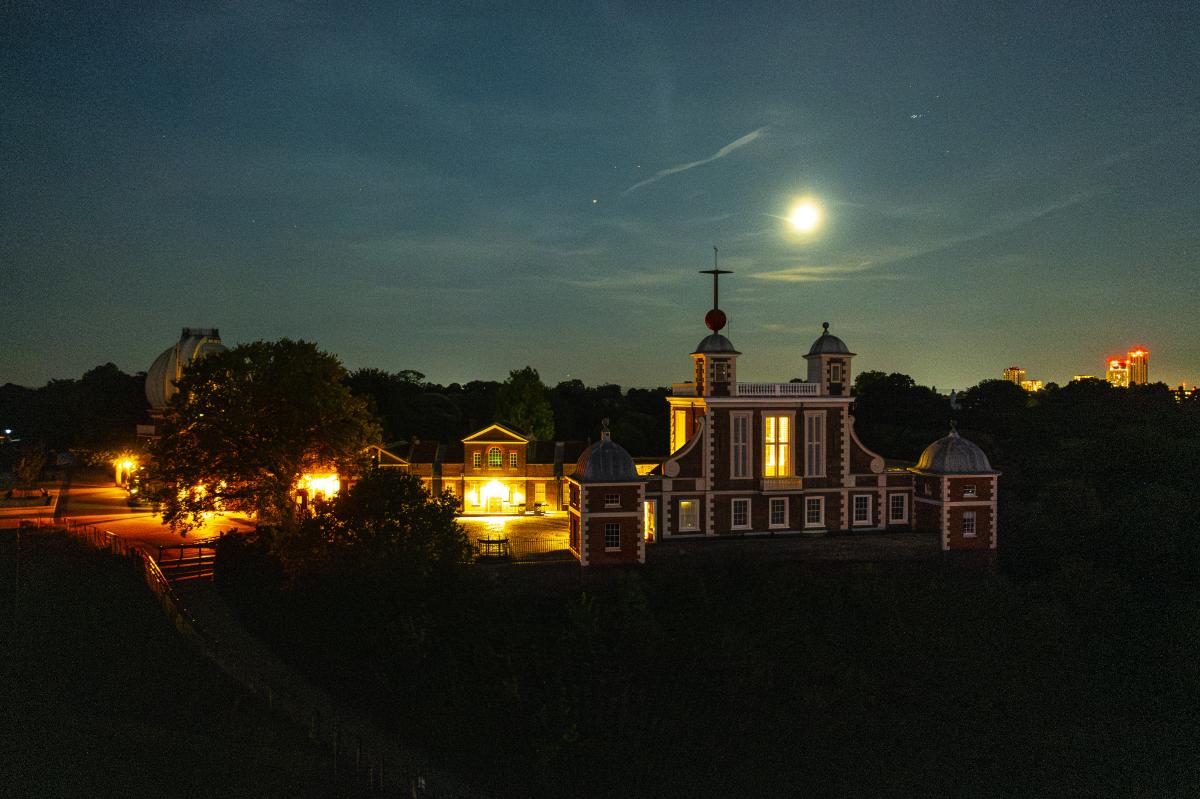 A drone photograph of the Royal Observatory at night, the Moon is bright in the sky and lights in the observatory buildings below casts a yellow glow