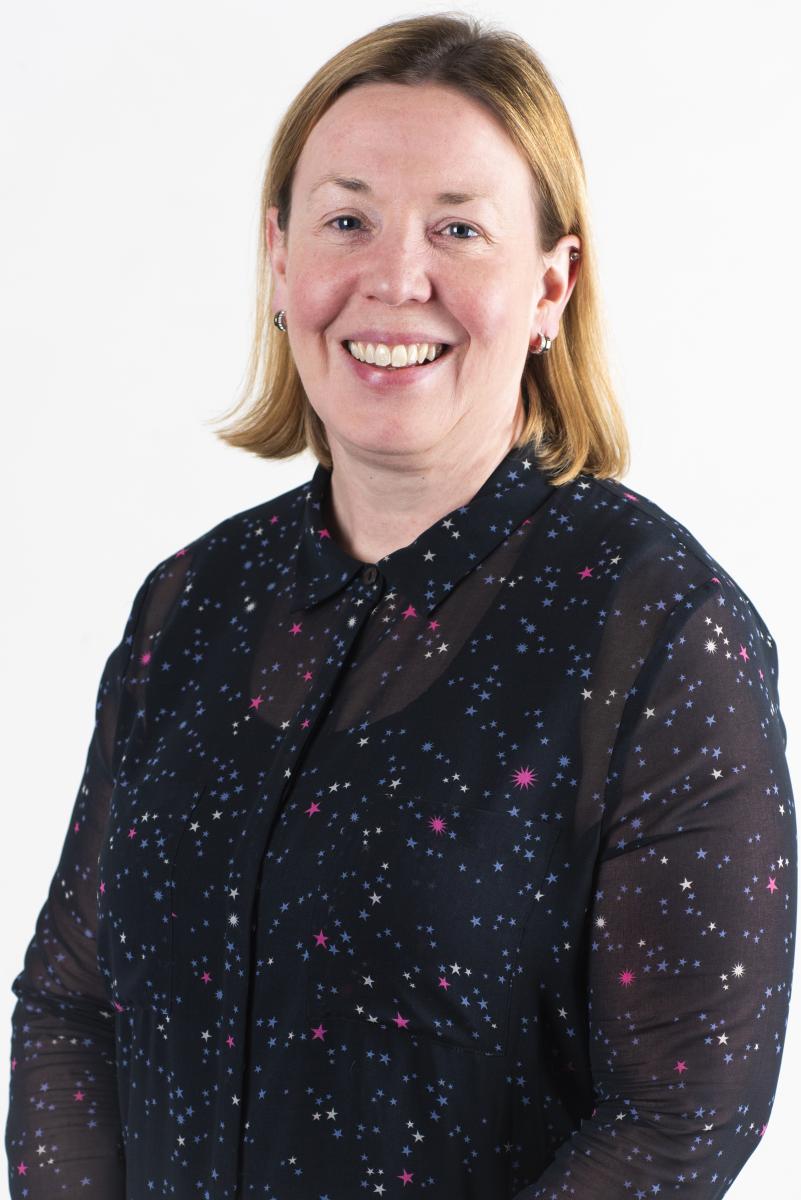 Portrait photograph of Professor Emma Bunch. She is smiling brightly at the camera, has light brown mid-length hair and is wearing a dark blouse with a pattern of bright pink, white and blue stars