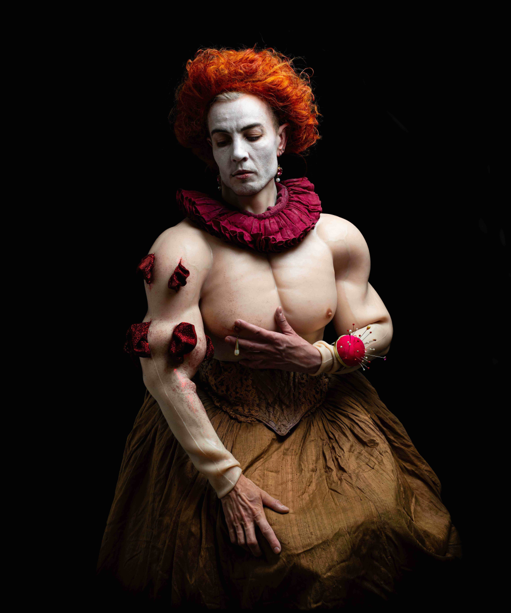 Artist E.M. Parry stands against a black background looking down over their shoulder. They wear a wide brown tudor skirt, a bear silicone torso, a red ruff around their neck and have a bright orange curly wig with white painted face