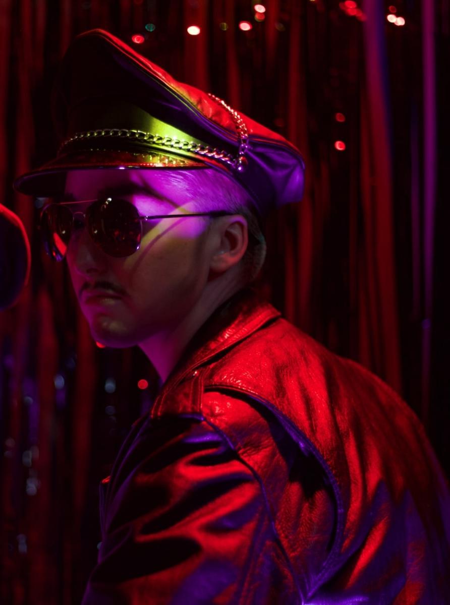 A darkly lit photograph of a person wearing a leather cap, dark glasses and leather jacket. They appear mysterious against a red and purple light and a plush curtain