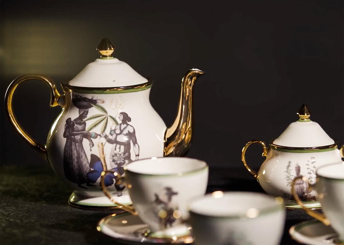 A delicate-looking tea service featuring teapot and matching cups. The central motif features a Black woman with a child on her back, handing an item to a bare-chested figure