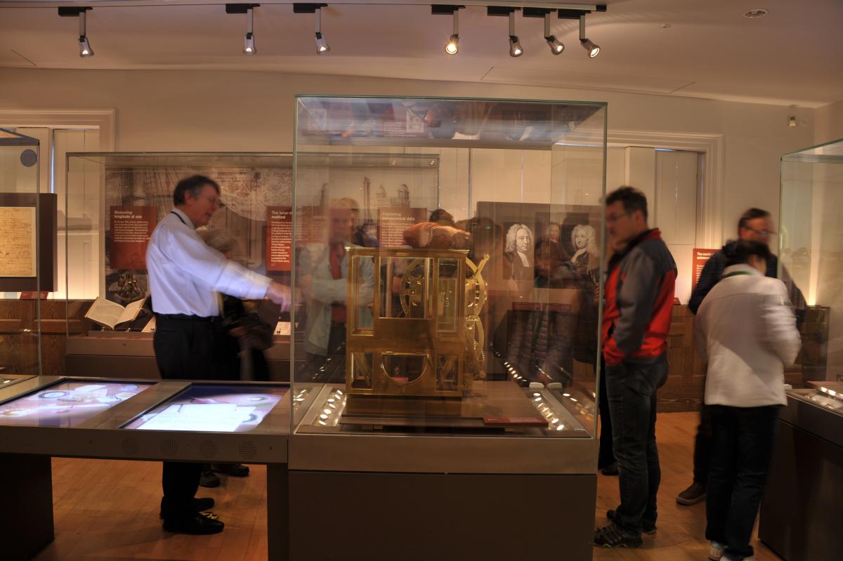 Photo of a man giving a tour in a museum, looking at a large golden clock, he is pointing at the object and a group of people are looking at it