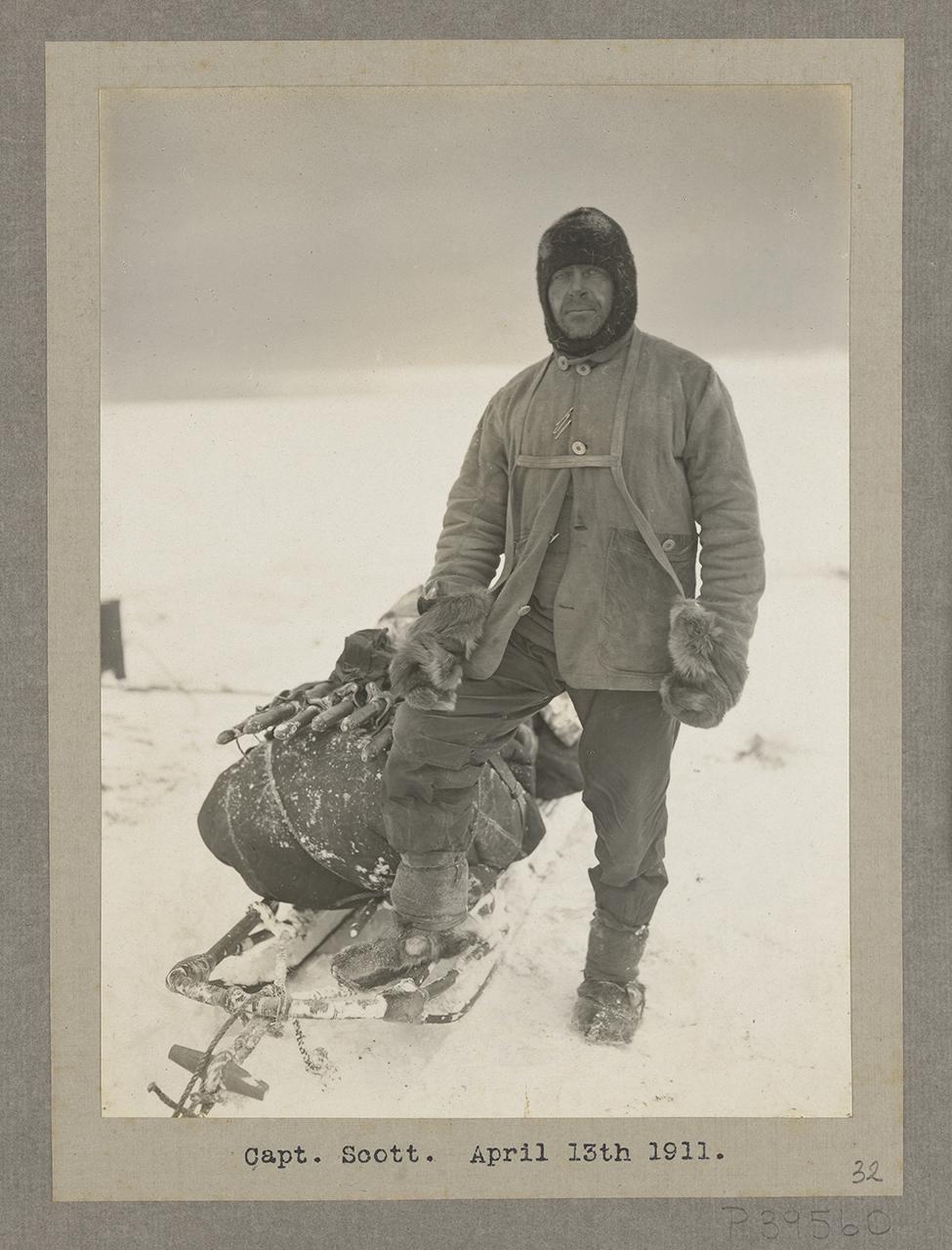 A full-length photograph of Captain R. F. Scott in full polar clothing standing next to the Nansen-designed sledge. His right foot is resting on the sledge frame with his right hand on his knee. The sledge is fully loaded with equipment and the tent poles can be seen lashed to the top. Scott is wearing a canvas over-jacket with the canvas webbing attached to large fur gloves, a knitted balaclava, and over-trousers tied with putties over finnesko fur boots. The photograph was taken on the day that Scott retu