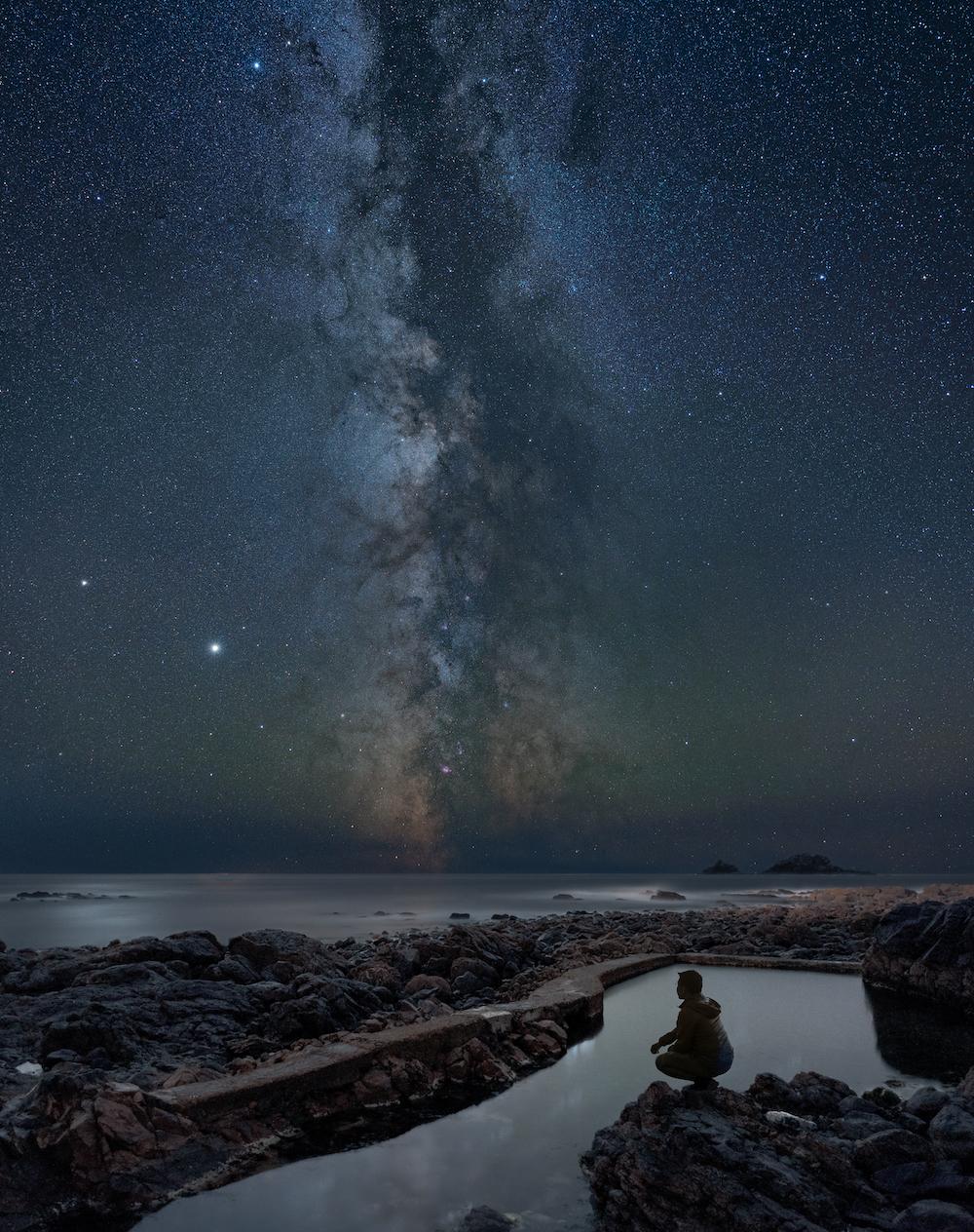 A person stares into a rockpool at night, with constellations soaring above