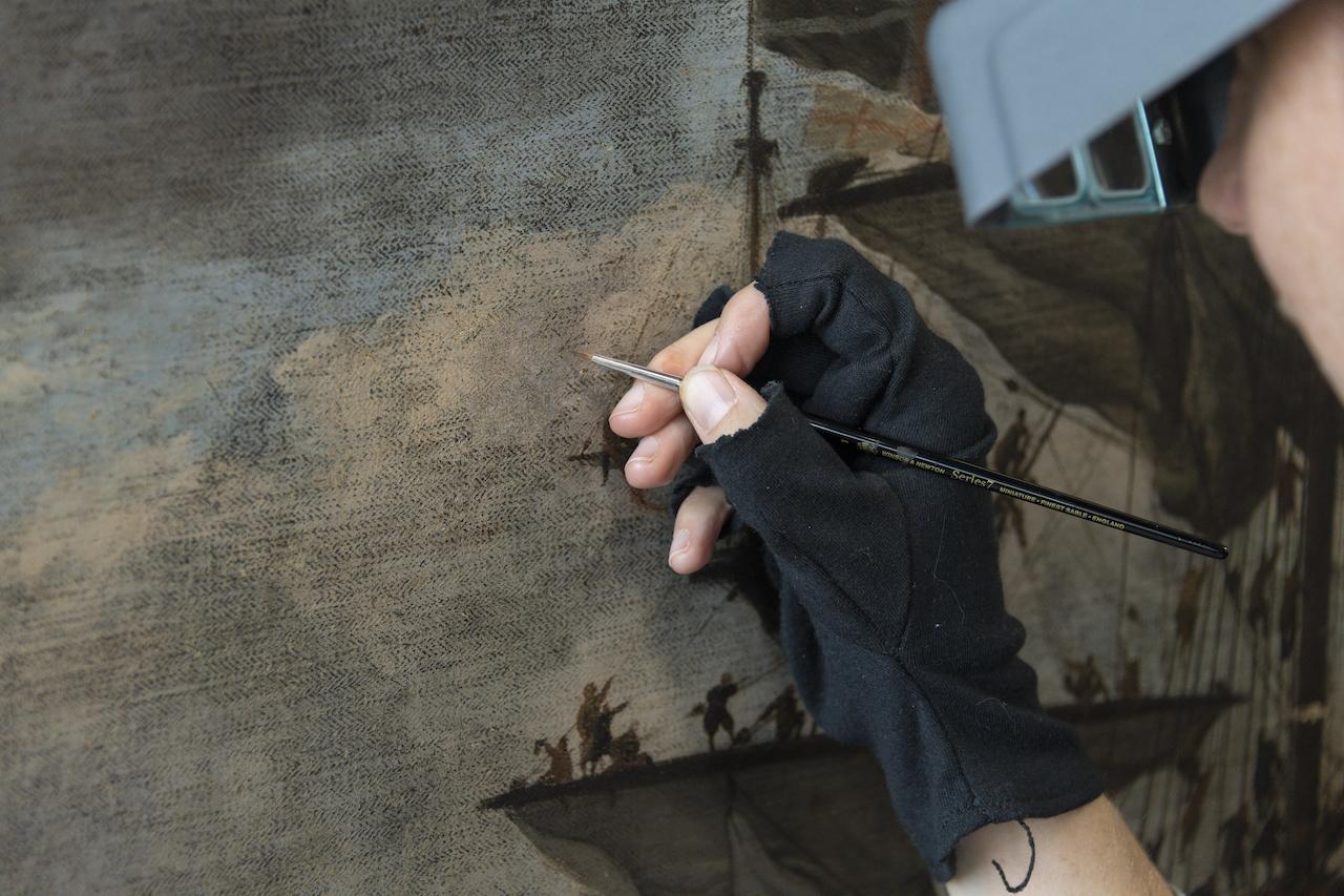 A paintings conservator carefully applies small amounts of retouching paint using a small brush and magnifying glasses