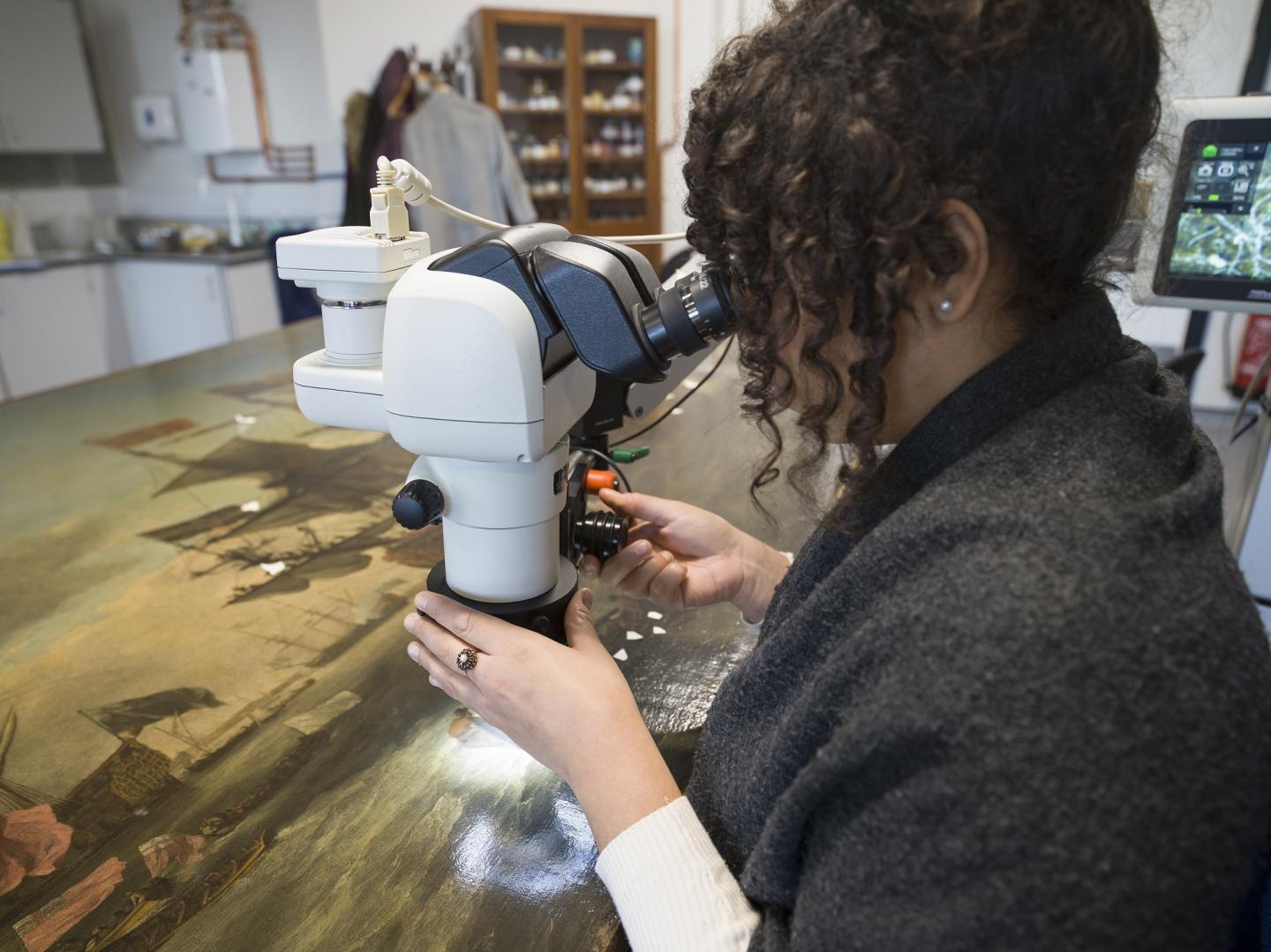 A woman analyses an oil painting using a microscope as part of the painting's conservation process