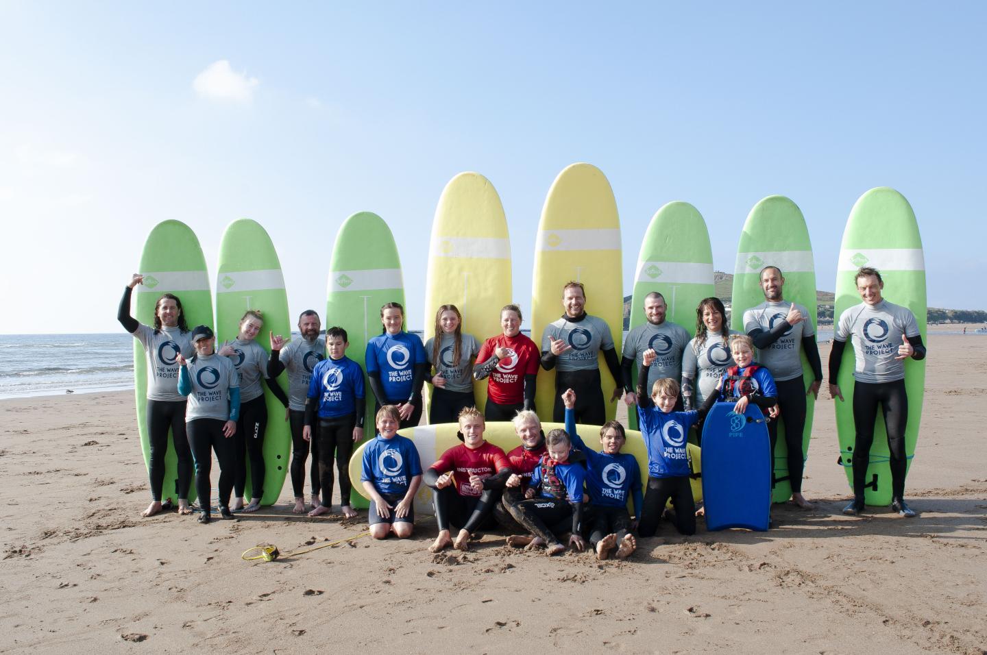 A group of people standing in front of surfboards with chidden sitting in front of them on a sandy beach. Blue sky in the background.