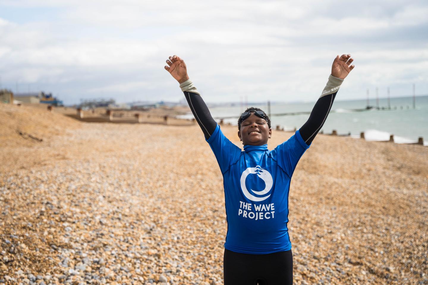 A young person with arms in the air, wearing a wetsuit and t-shirt, standing on a beach.