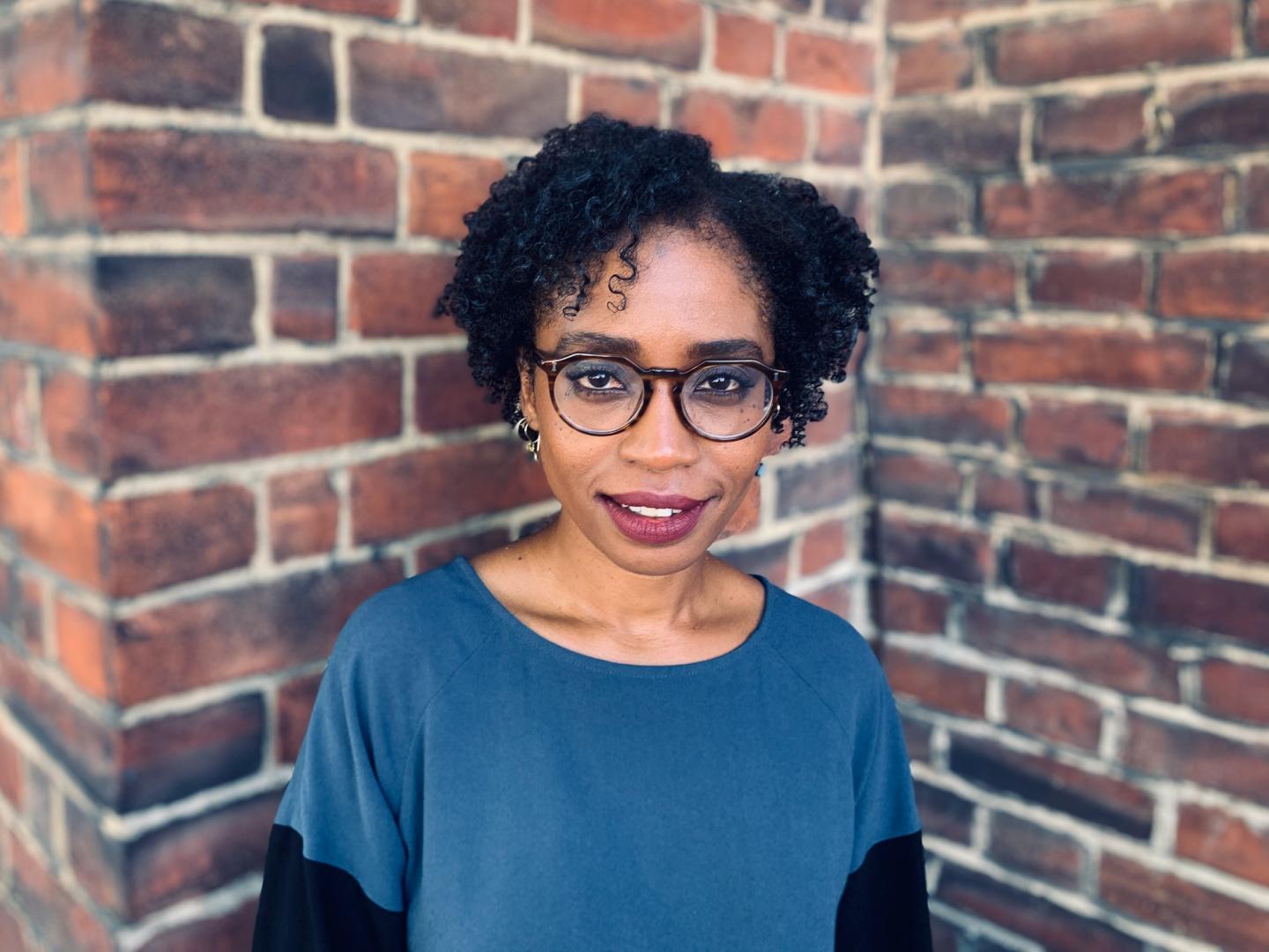 A headshot of Dr Nydia Swaby. Nydia is a Black woman with short black hair, wearing large glasses and a blue t-shirt standing in front of a brick wall. 