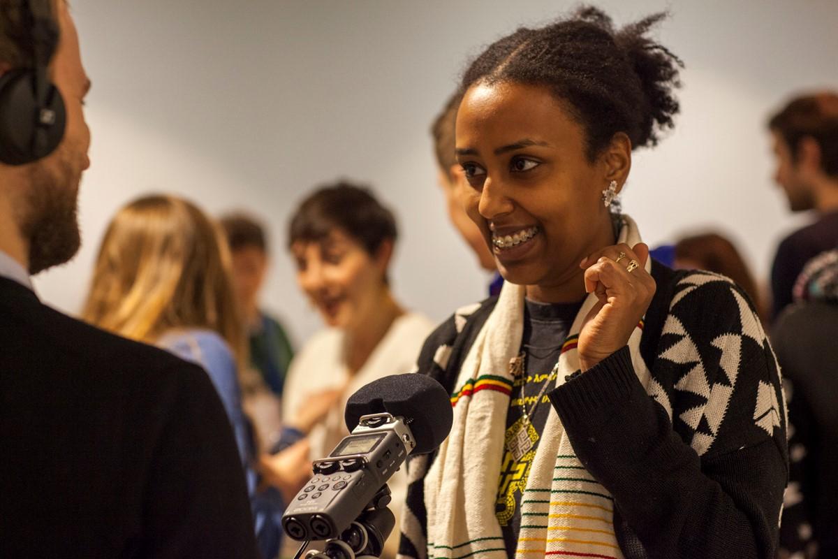 photo of a black woman smiling and facing a man who is interviewing her, with some people in the background