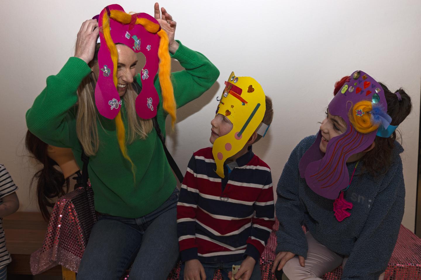 Photo of mother and two children showing each other their crafted pink, yellow and purple wigs