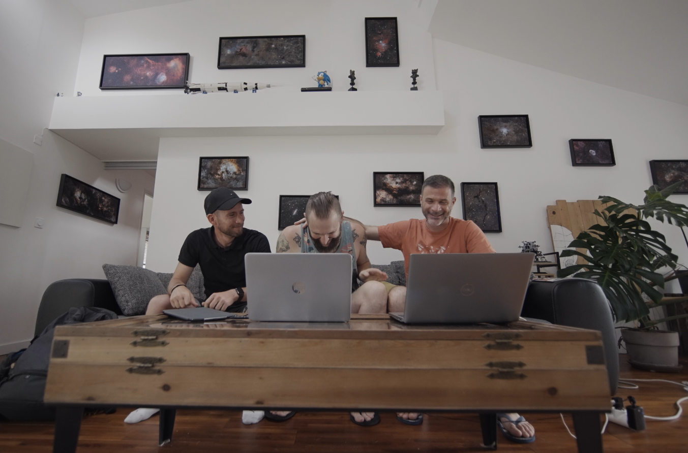 Image of three men sitting on a sofa with coffee table in front with laptops on it, the three men are laughing