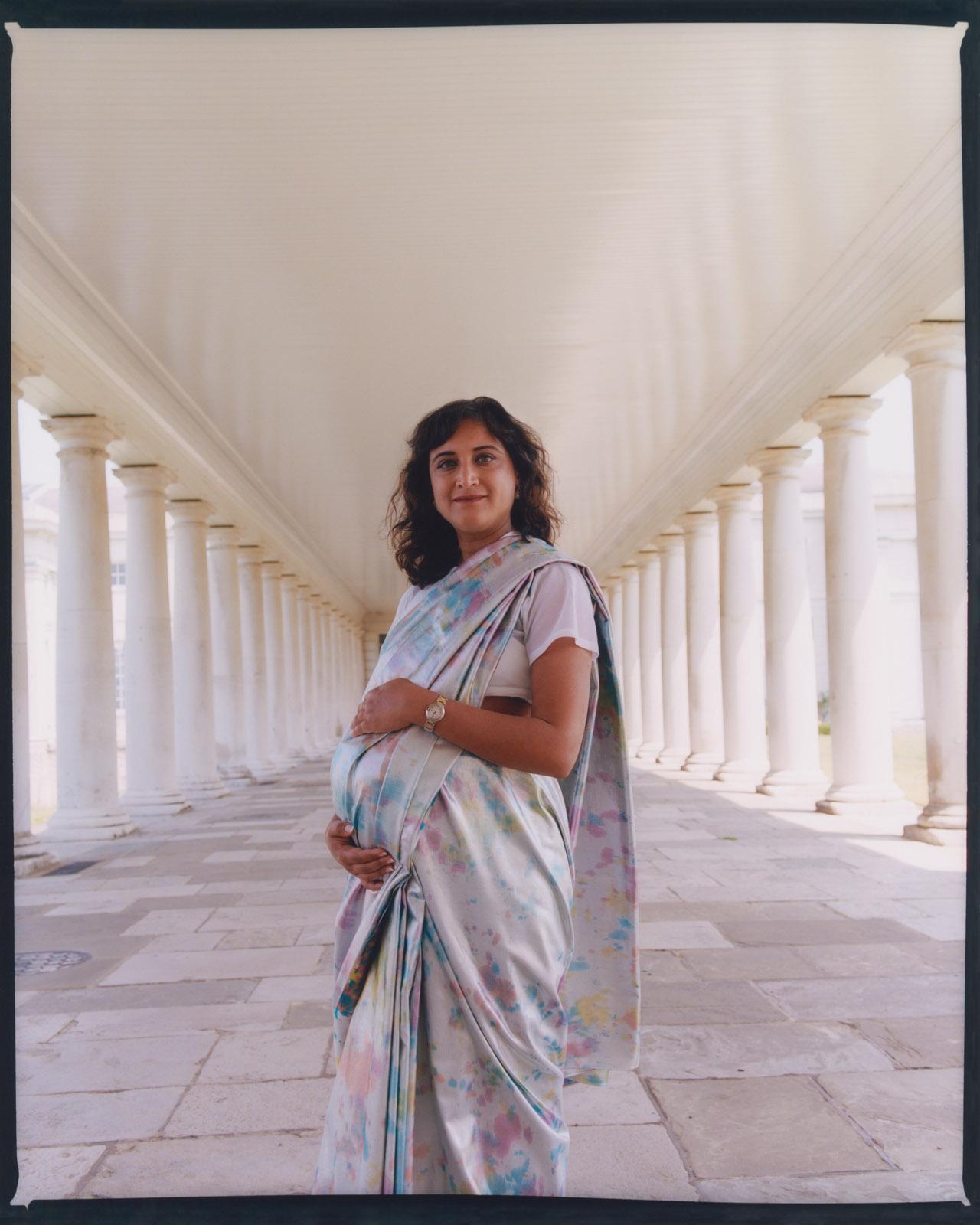 Portrait photograph of a pregnant woman wearing a white sari with light speckled pattern. She is standing right in the centre of a series of colonnades, perfectly framed by the white pillars on either side