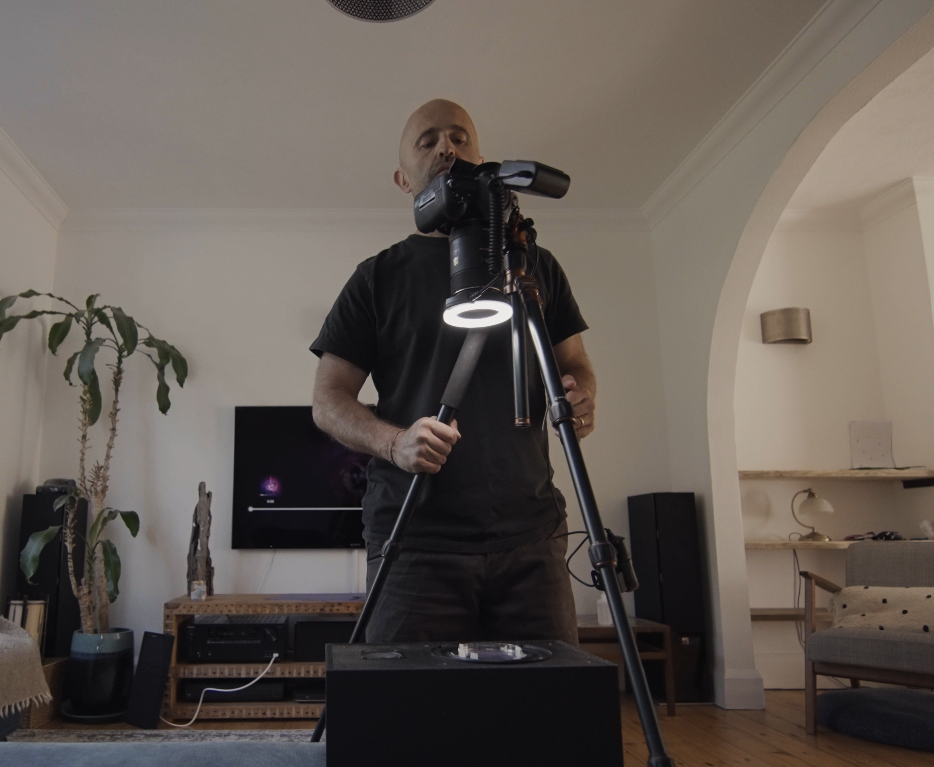 Image of man with a camera pointed downwards in a living room, pointing at a petri dish with water in it on top of a speaker