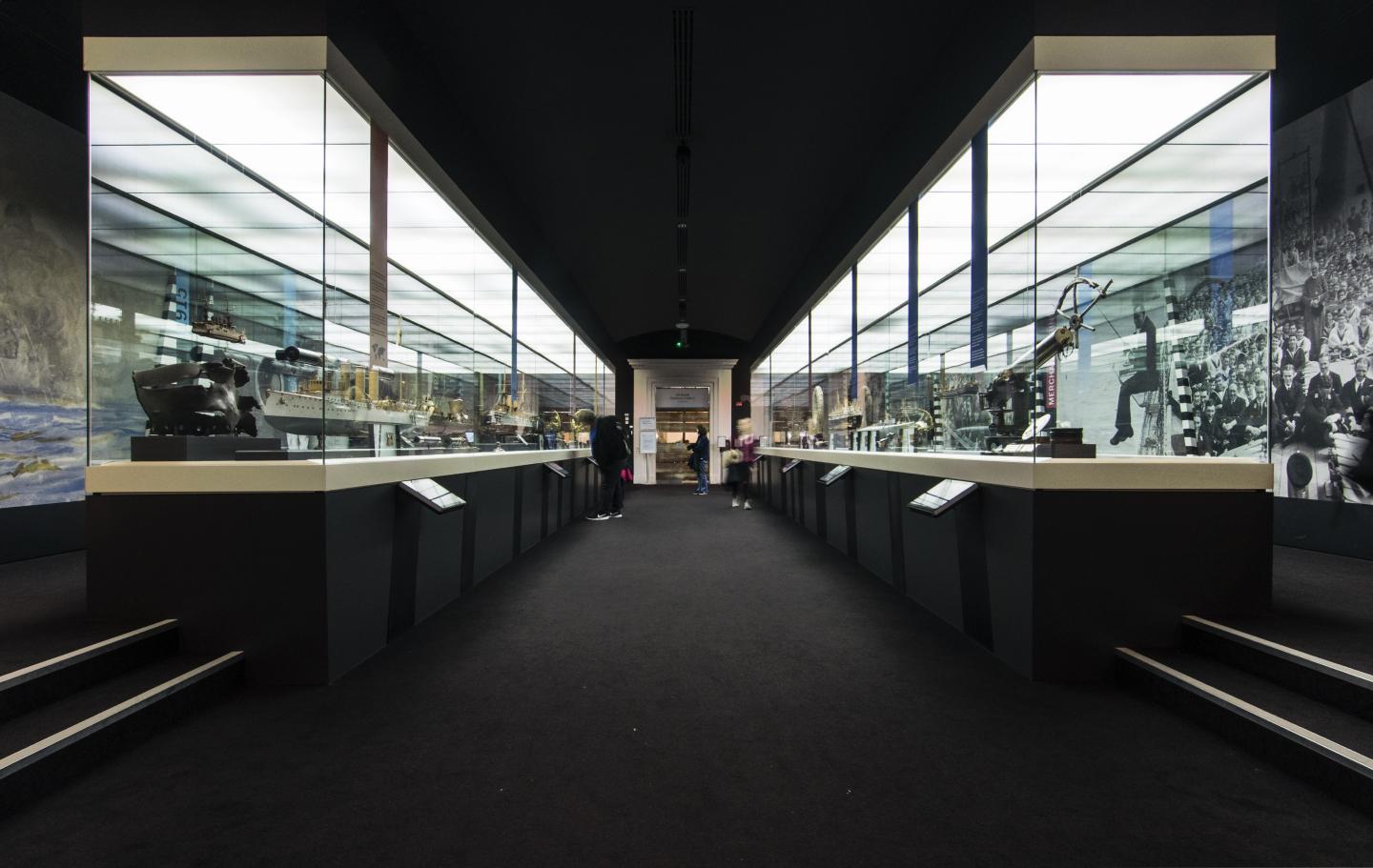 Image of the Forgotten Fighters gallery which has a walkway in the middle of two big glass cases that span the length of the room. They are filled with objects relating to the first world war at sea