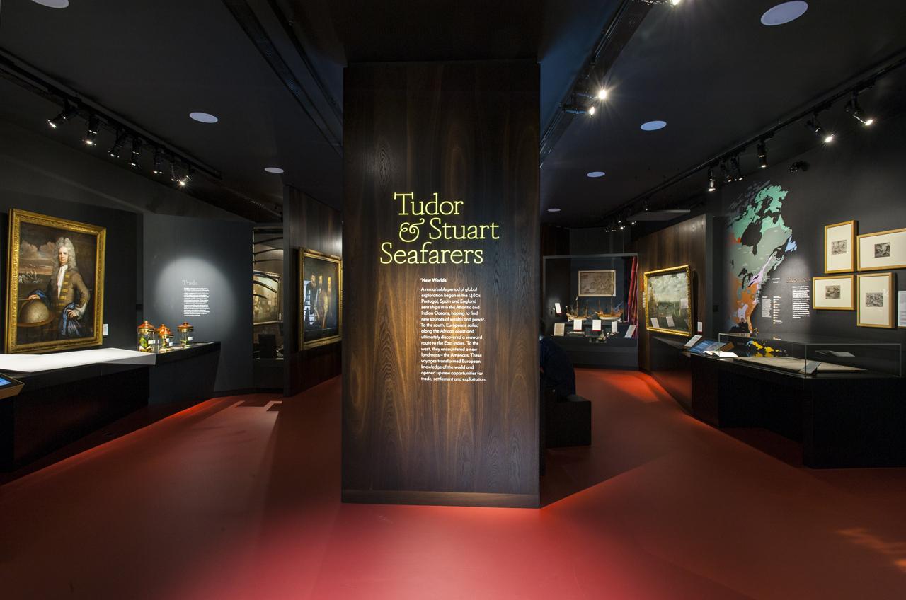 Photo of the Tudor and Stuart seafarers gallery at the National Maritime Museum, with displays and exhibits along the walls and on stands in the middle of the room. The floor is red and there are many paintings lining the walls, with spotlights on the ceiling