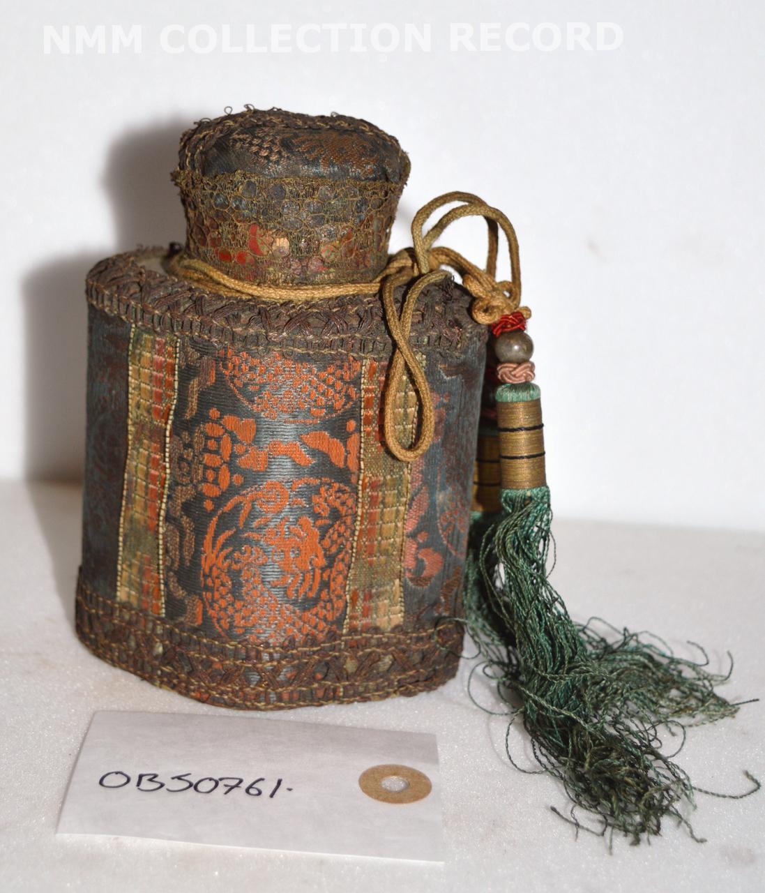 Metal tea caddy with a lid, covered in Chinese embroidery of a type known as 'a present'. The caddy is shaped and the damask and embroidered panels are edged with gold lace. It is secured with two green silk beaded tassels.