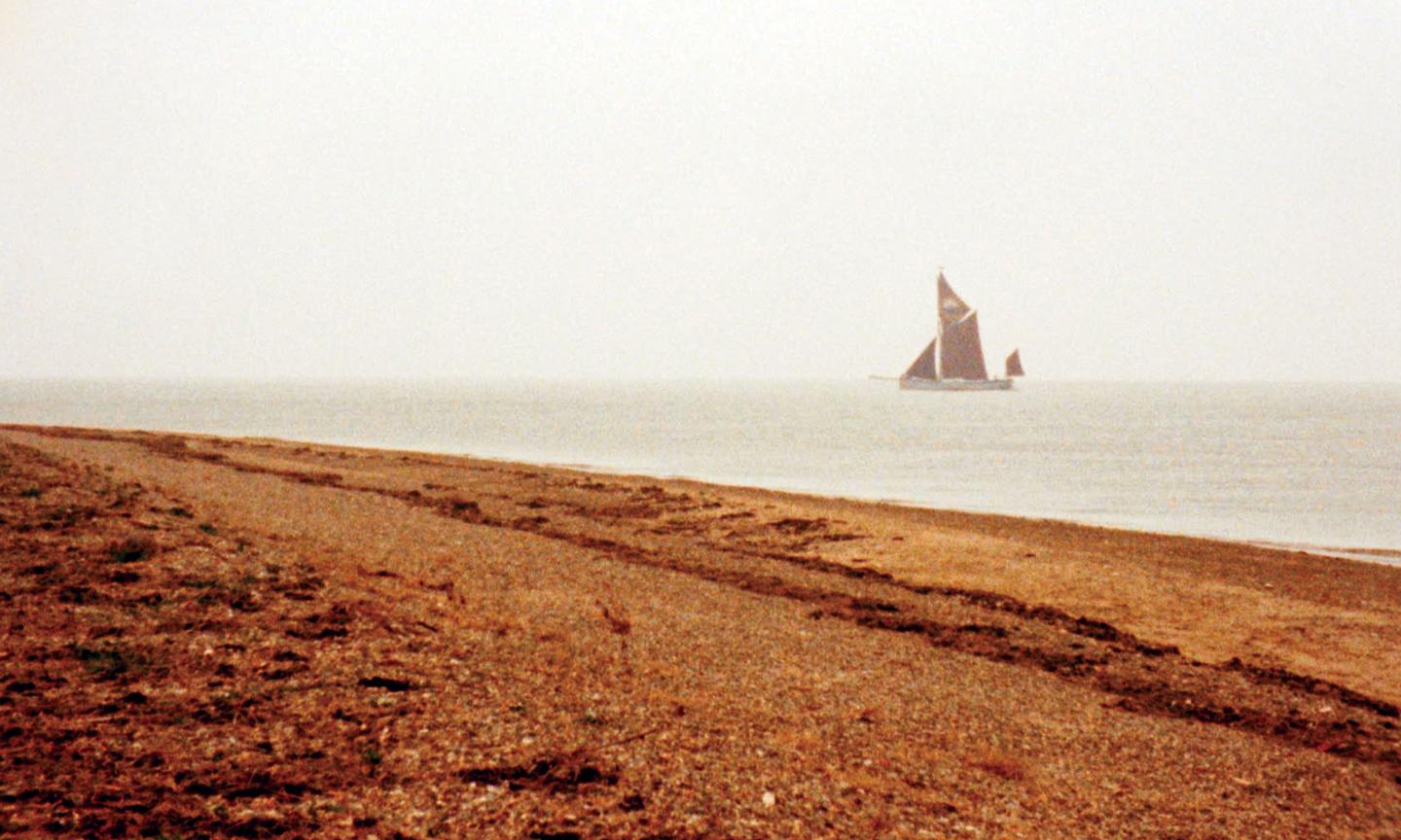The foreground is a long brown beach with a vessel Thames sailing barge Blue Mermaid sailing in the distance, with four red sails. 