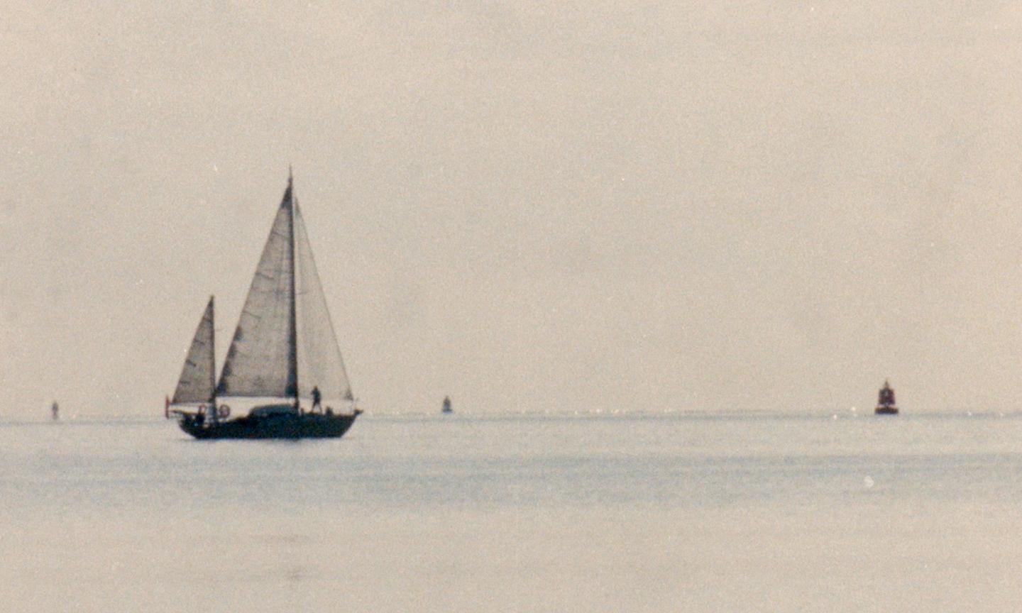 The sky and water are a washed out pale colour. The foreground is of a small sailing vessel with three white sails flown and the silhouette of a person on deck. There are navigation markers visible.