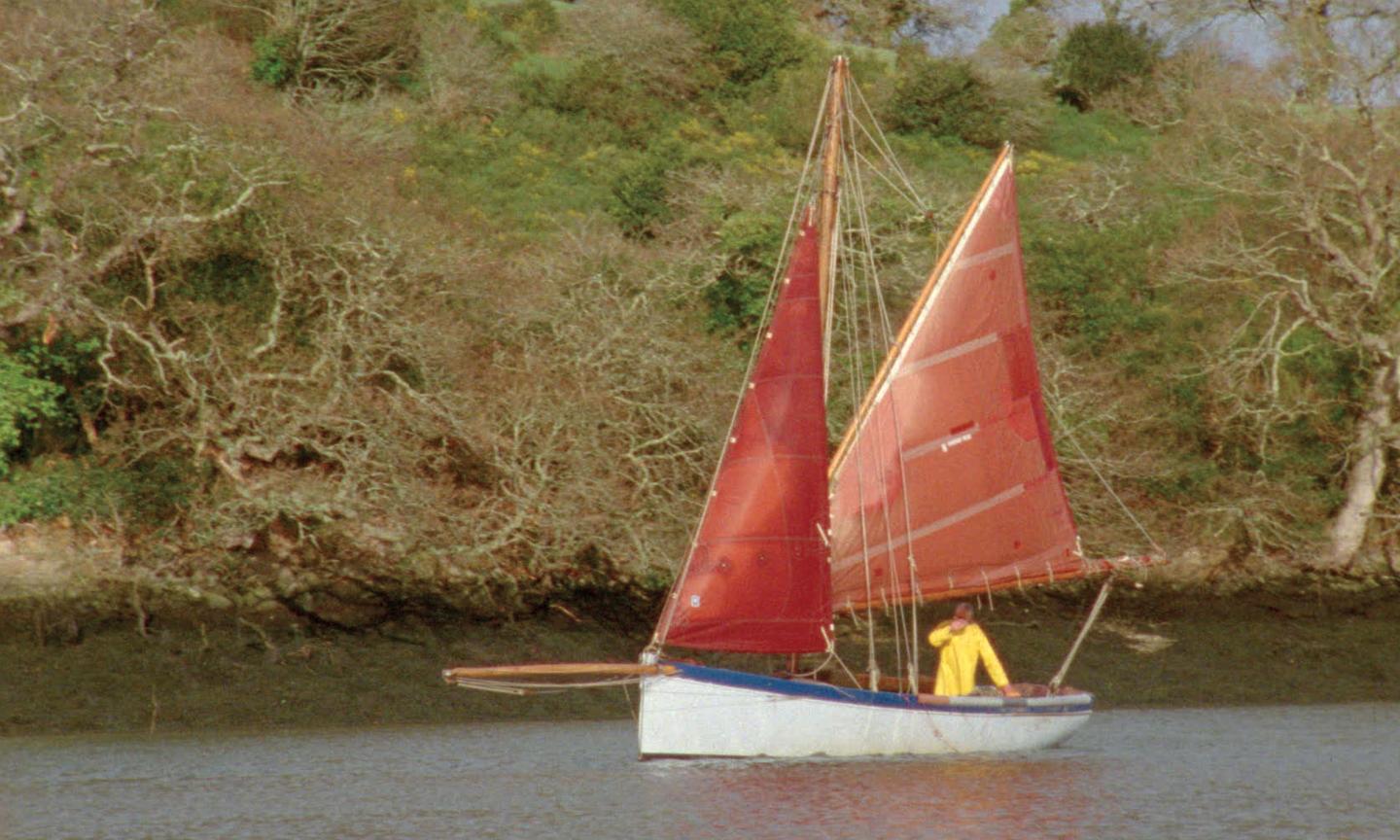 A small white sailing boat with a blue rim and two red sails is in the foreground, behind the boat are trees and a riverbank. On board the boat is a man in a yellow coat.