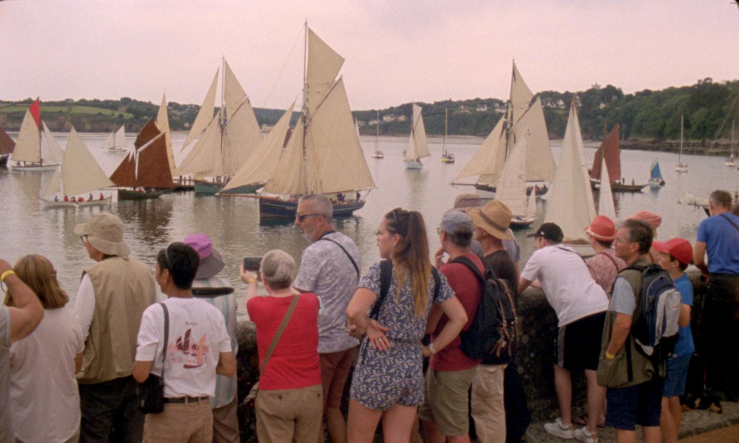 In the foreground, a crowd of people on land are looking out onto a number of small sailing vessels, with their sails up, a mix of red and white. There are hills, trees and fields in the background.