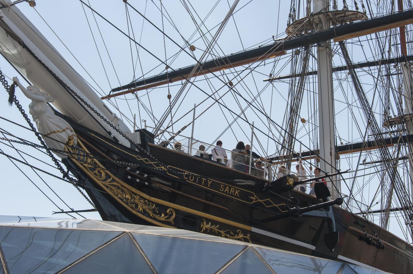 A view from below of an old sailing ship on a glass platform, with a long bow under which is a white figurehead in the shape of a woman. There are lots of ropes which form the rigging zigzagging across the image.