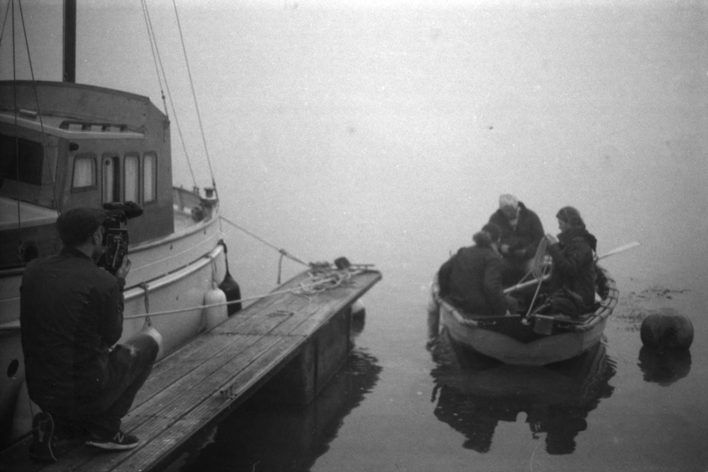 A man on a jetty holding a film camera, filming three people in a small rowing boat, leaving the jetty.
