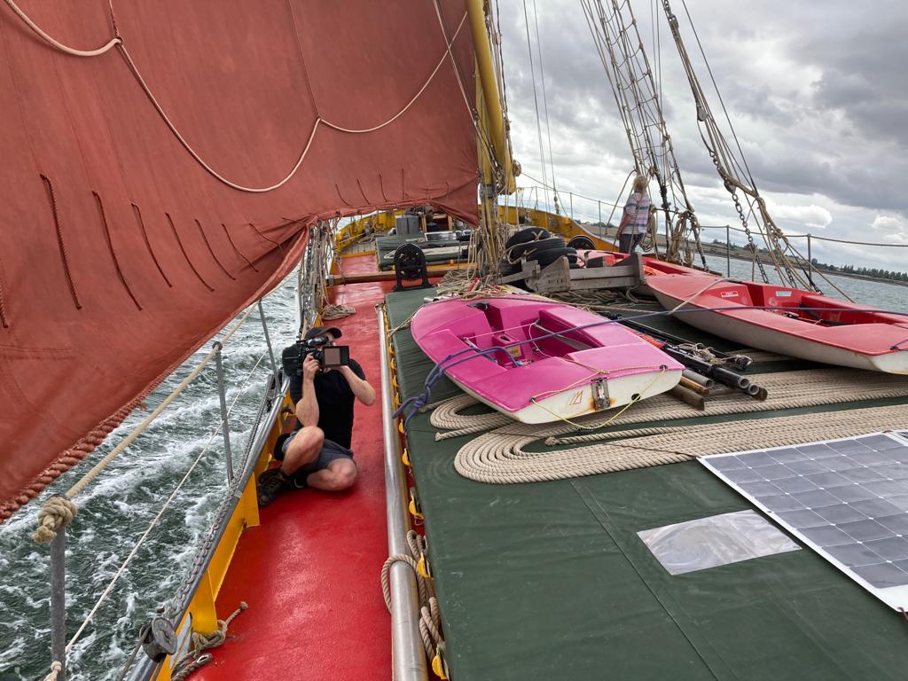 A man holding a camera sits on one side of a sailing vessel, Thames barge Blue Mermaid, while it is sailing, using a large red sail, filming towards the back of the vessel. 